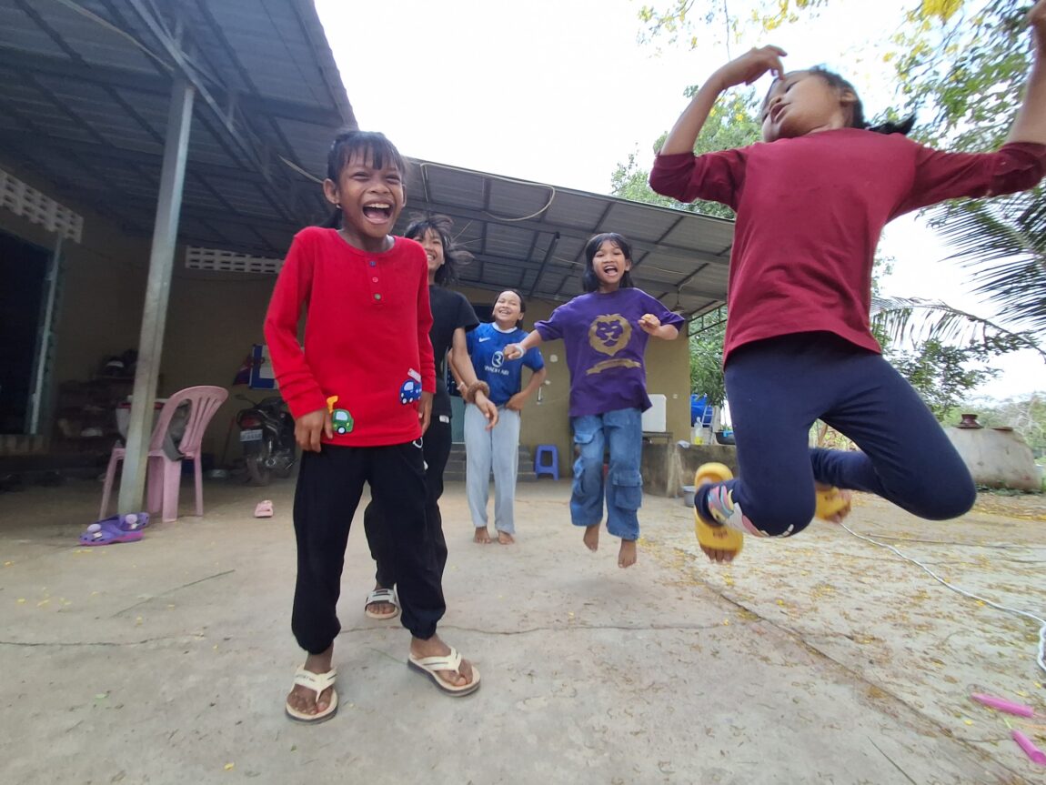 Children laughing and jumping in an outdoor courtyard; a girl in a red shirt stands in the foreground while others behind her join the fun.