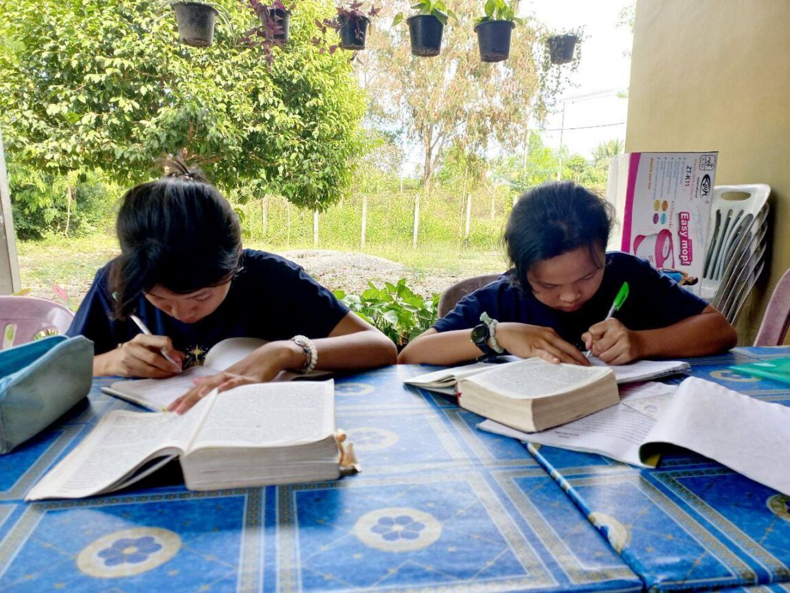 Two young girls study at a blue patterned table outdoors, writing in notebooks with open books and pencils, surrounded by plants and trees on a sunny day.