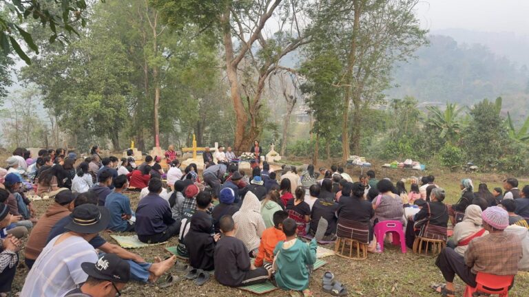 Large outdoor ceremony with a speaker at a podium addressing a crowd seated on mats under trees in a rural setting.",
