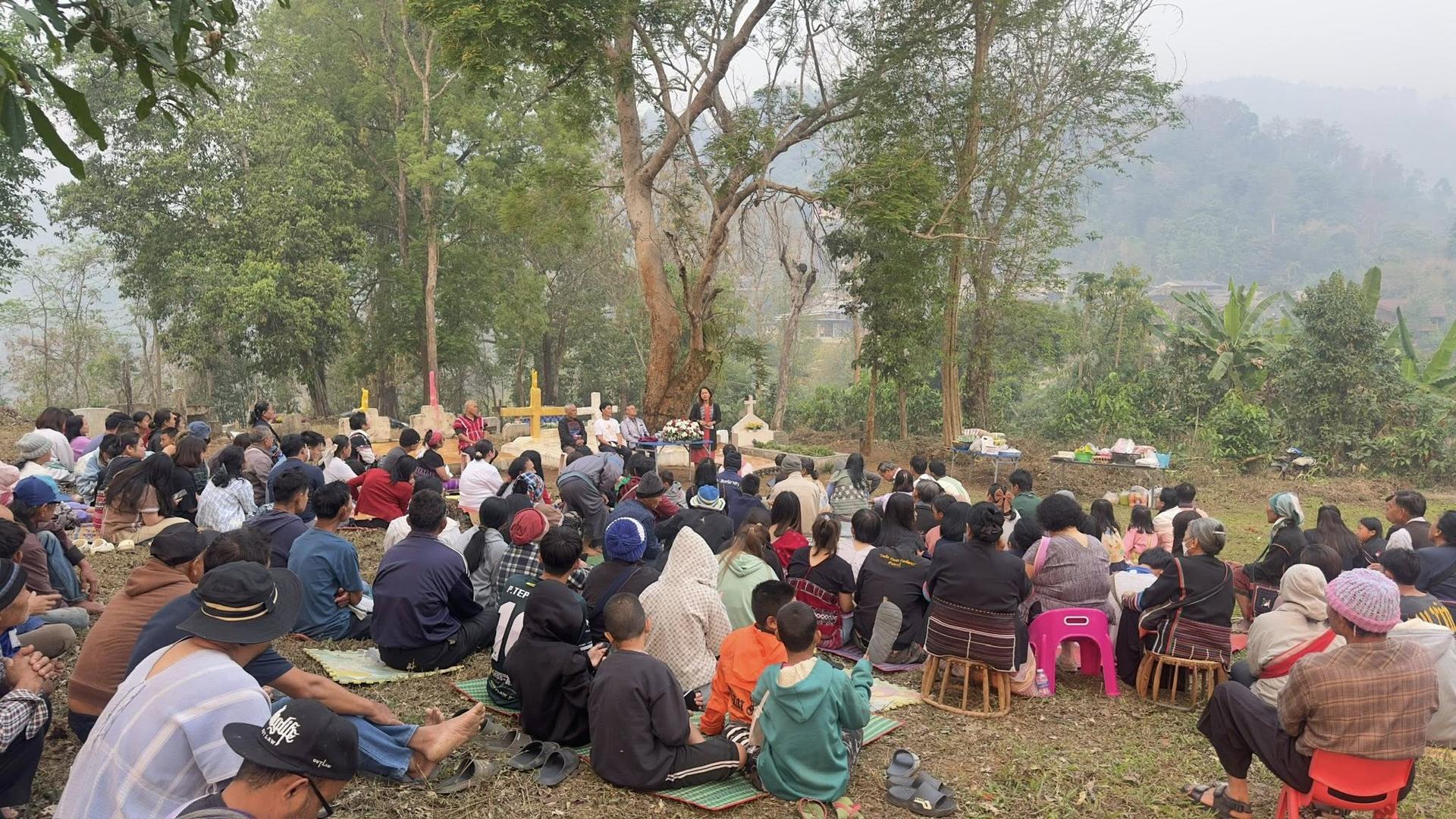 Large outdoor ceremony with a speaker at a podium addressing a crowd seated on mats under trees in a rural setting.",